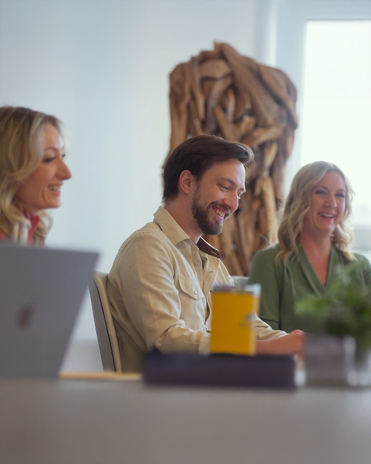 Three colleagues laughing together during a meeting, with a laptop and drinks on the table and a large wood sculpture in the background.