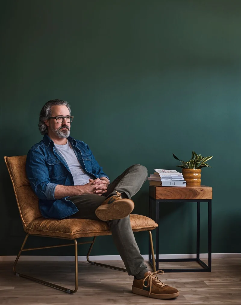 A middle-aged man with glasses sitting relaxed in a leather chair against a dark green wall, with a side table holding books and a plant beside him.
