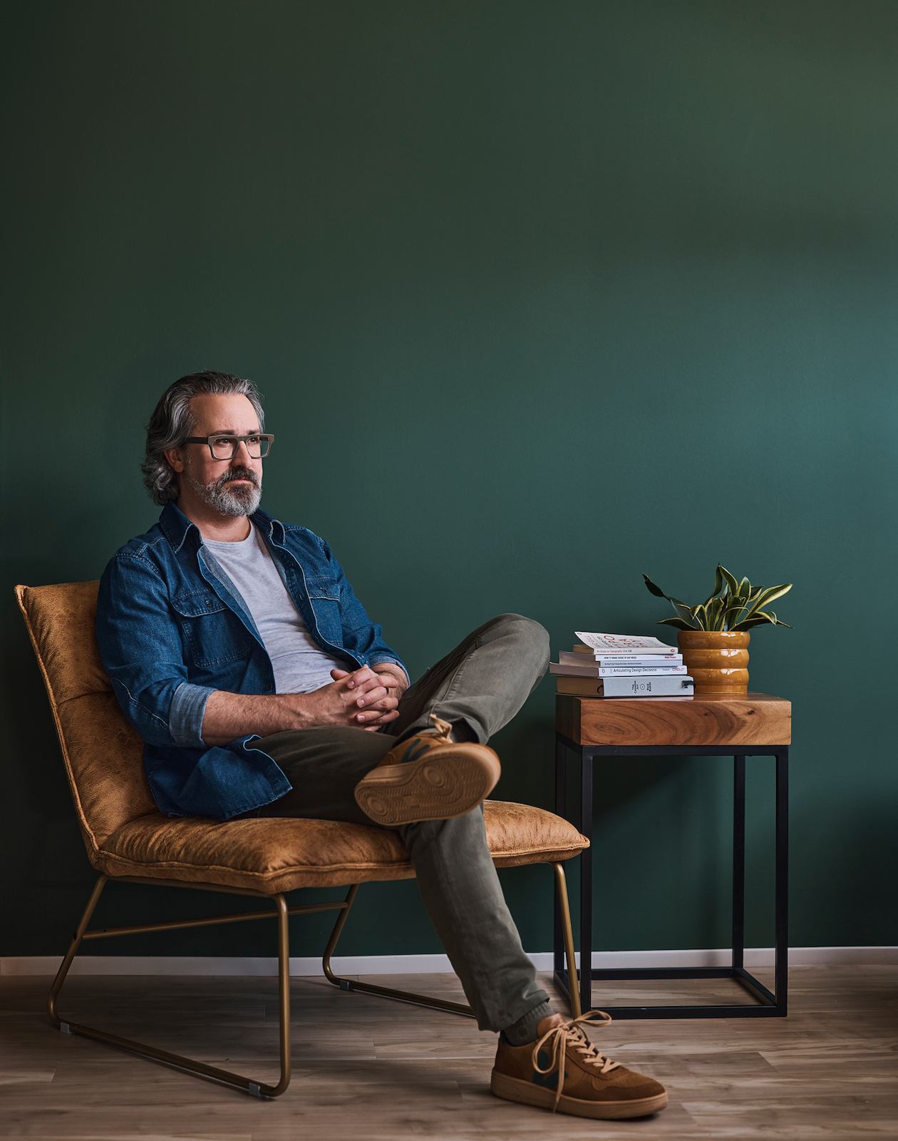 A middle-aged man with glasses sitting relaxed in a leather chair against a dark green wall, with a side table holding books and a plant beside him.