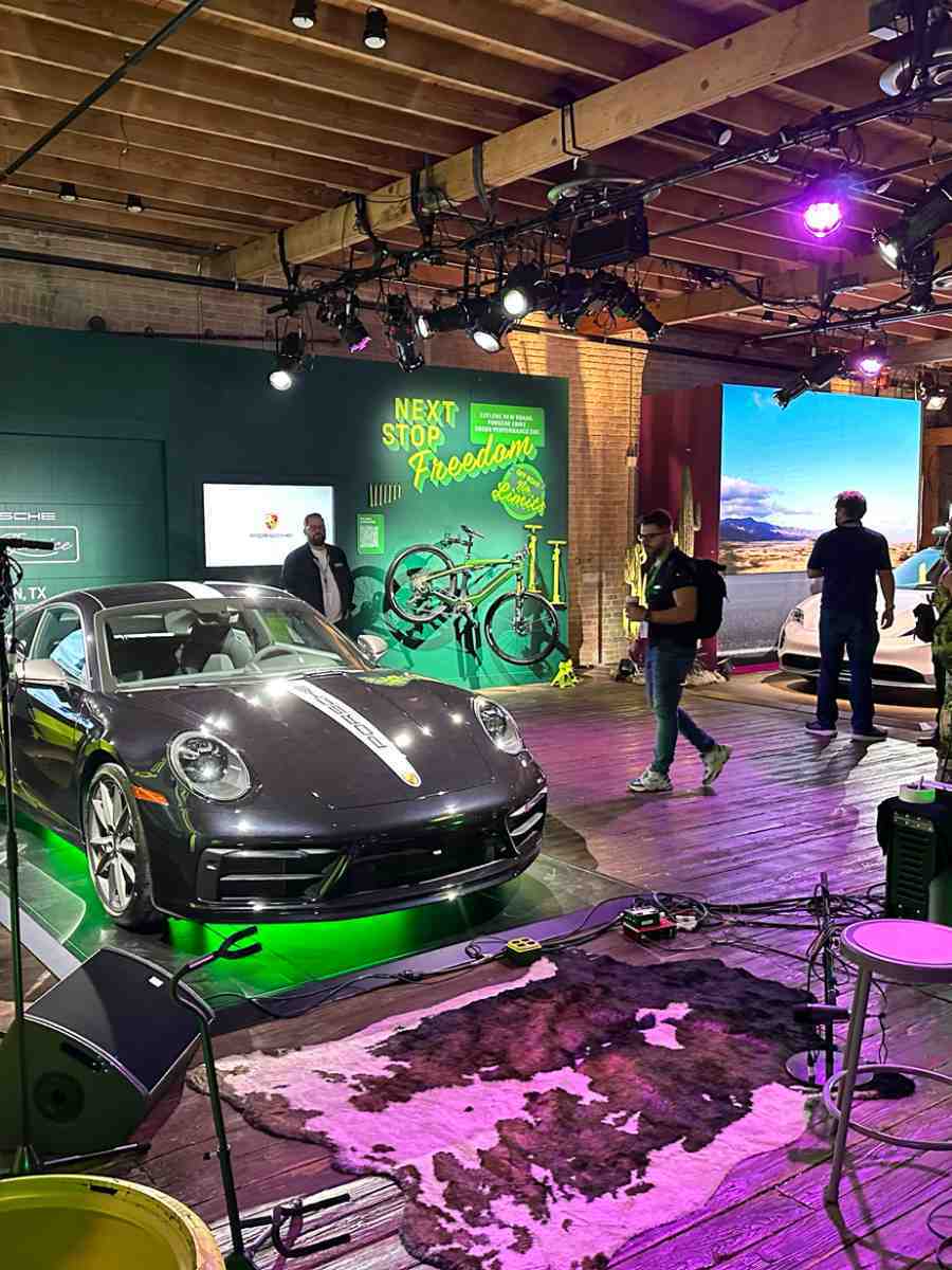 A Porsche 911 on display at an indoor event space with purple stage lighting, bicycles, and a "Next Stop Freedom" branded backdrop.