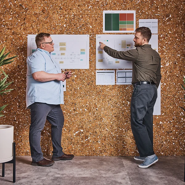 Two men standing in front of a cork wall reviewing project planning boards with sticky notes, charts, and timelines pinned to them.