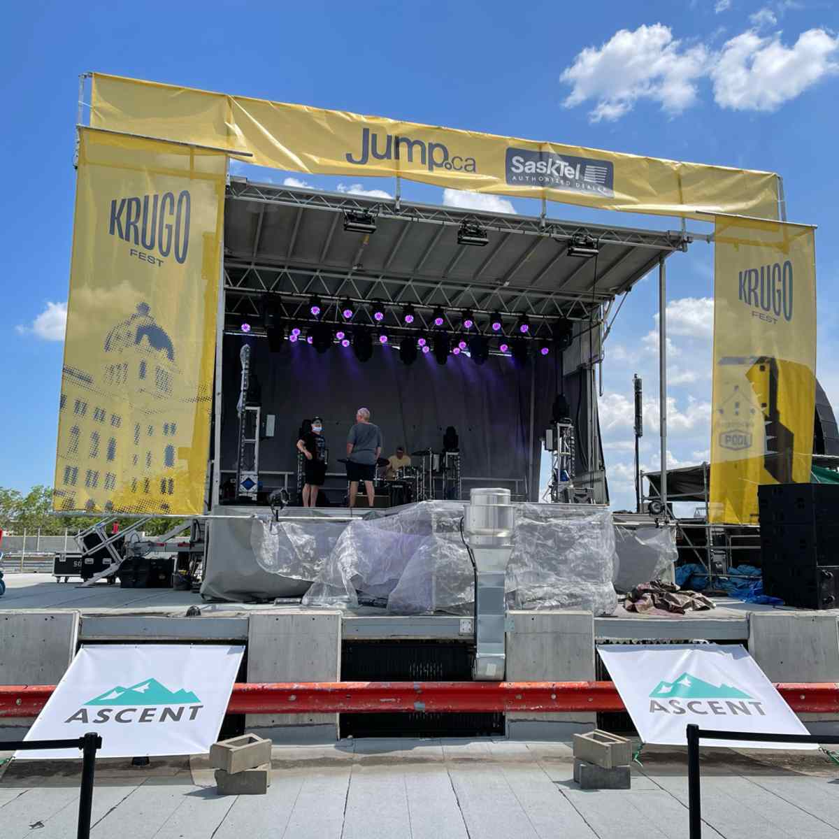 An outdoor concert stage branded with Jump.ca, SaskTel, Krugo, and Ascent signage being set up under a blue sky.