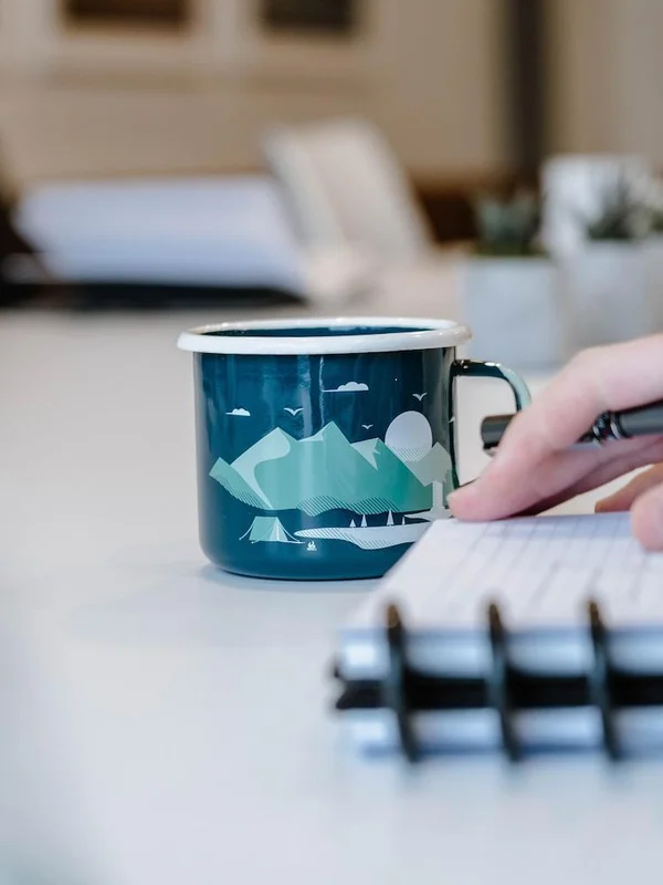 A teal enamel camp mug with a mountain landscape design on a desk beside a hand writing in a notebook.