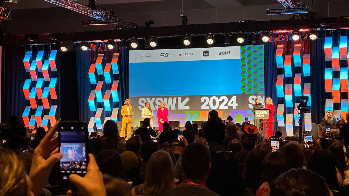 Speakers on stage at SXSW 2024 in front of a large screen and colorful geometric backdrop, with a packed audience in the foreground.