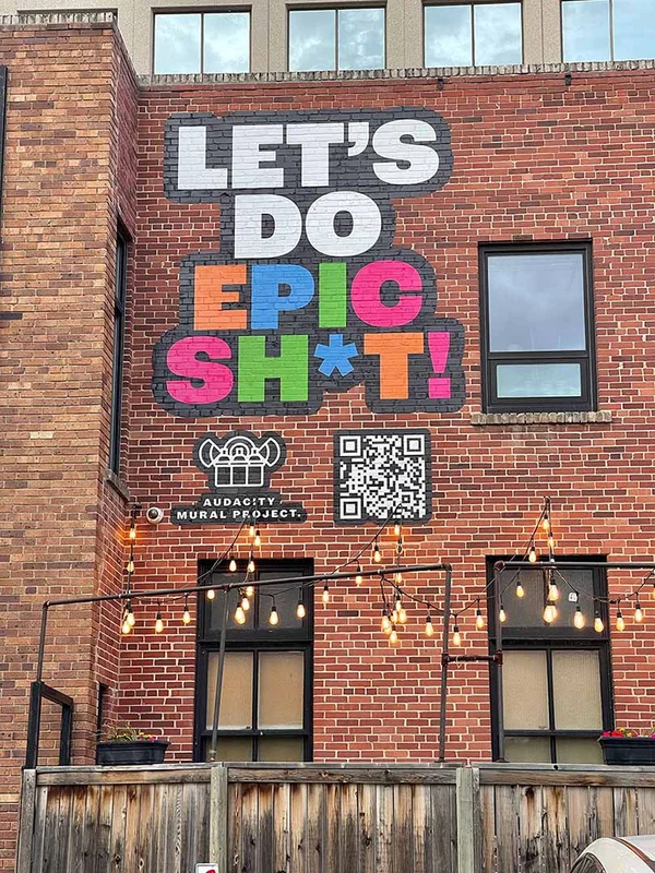 brick building with a large mural painted on its side. The mural features the phrase 'LET'S DO EPIC SH*T!' in bold, colourful letters