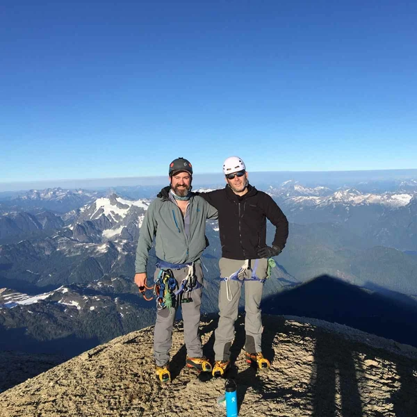 Two mountaineers in climbing gear posing on a rocky summit with a vast mountain range and clear blue sky behind them.