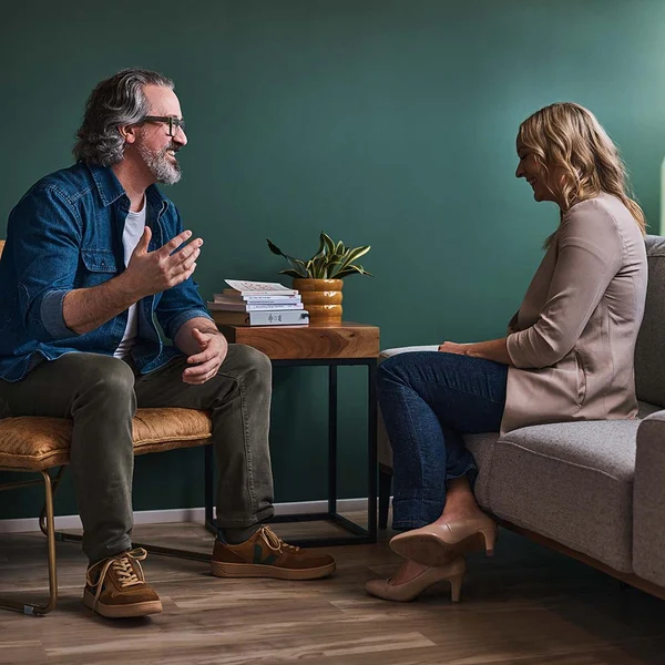 Jeff and Nicole sitting across from each other at a small wooden coffee table in a room with green walls. He is gesturing with his hands as if he is speaking or explaining something.