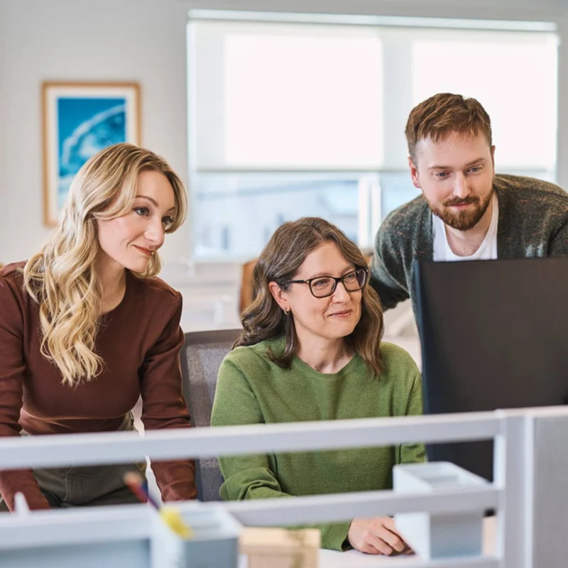 Three coworkers gathered around a computer monitor in an office, collaborating on work together.