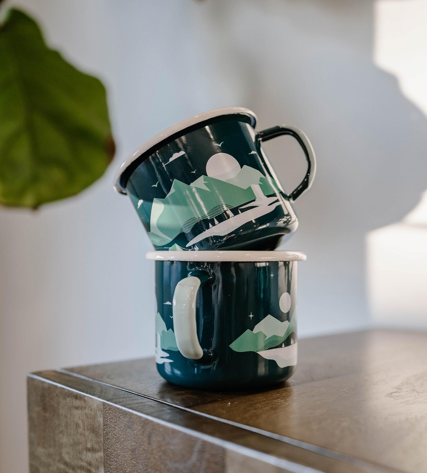 Two teal enamel mugs with an abstract green and white pattern stacked on a wooden surface.