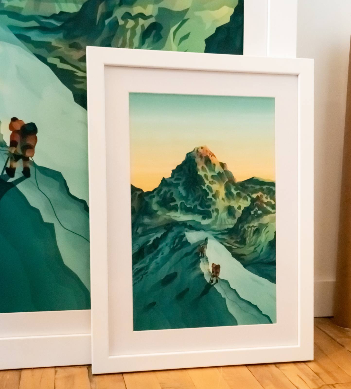 Two framed prints leaning against a wall, depicting illustrated mountaineers climbing a dramatic snow-covered peak at sunset.