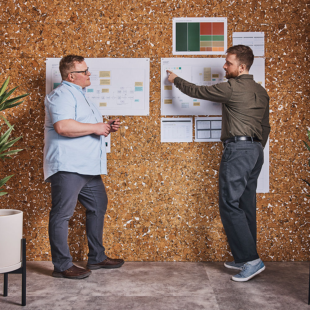 Two men standing in front of a cork wall reviewing project planning boards with sticky notes, charts, and timelines pinned to them.