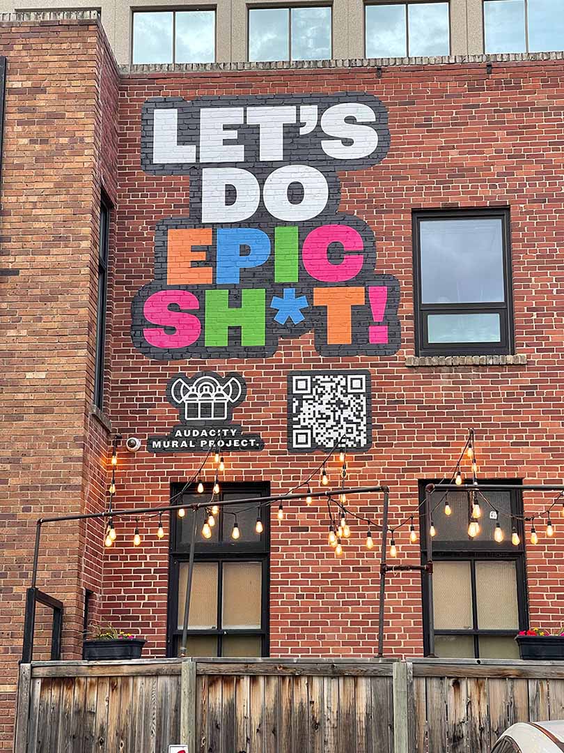 brick building with a large mural painted on its side. The mural features the phrase 'LET'S DO EPIC SH*T!' in bold, colourful letters