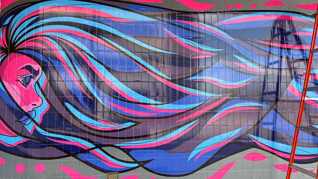 Street mural on a brick wall depicting a woman's face in profile with long, flowing hair in shades of pink and blue.