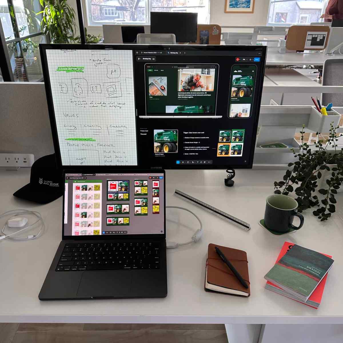 A dual-monitor desk setup showing website design work, with notebooks, a coffee mug, and a whiteboard with wireframe sketches nearby.