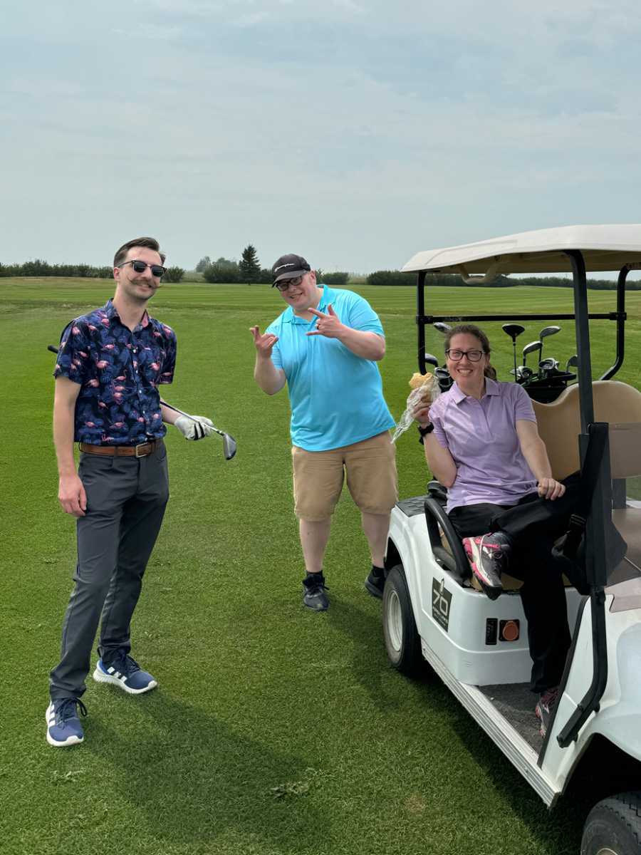 Three people posing and smiling on a golf course beside a golf cart on a sunny day.