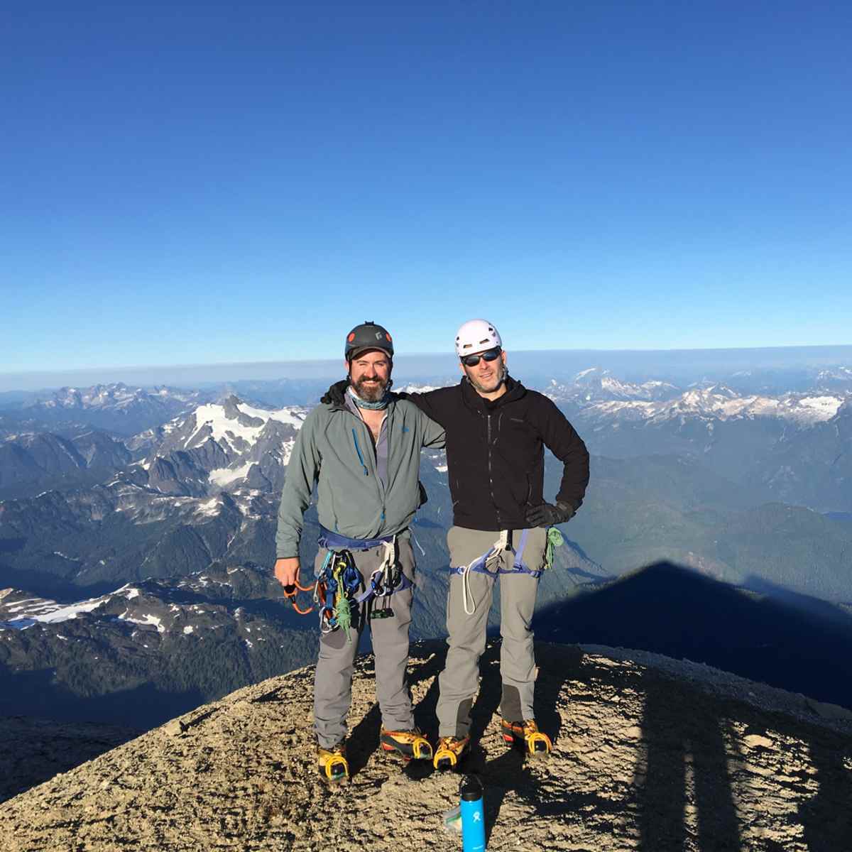 Two mountaineers in climbing gear posing on a rocky summit with a vast mountain range and clear blue sky behind them.