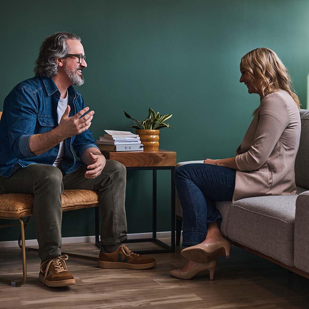 Jeff and Nicole sitting across from each other at a small wooden coffee table in a room with green walls. He is gesturing with his hands as if he is speaking or explaining something.