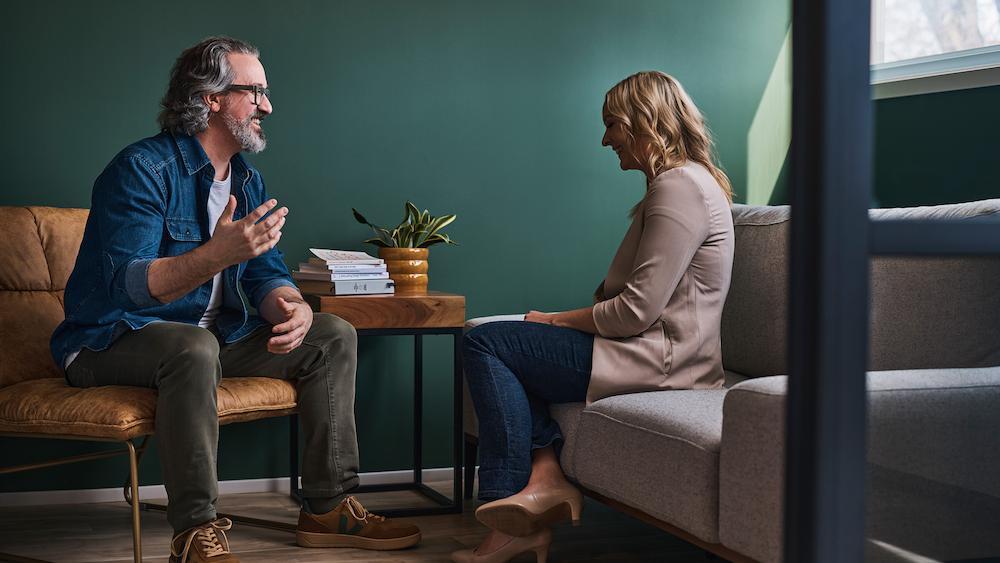 Two people engaged in conversation while seated across from each other in a modern lounge area with teal walls.