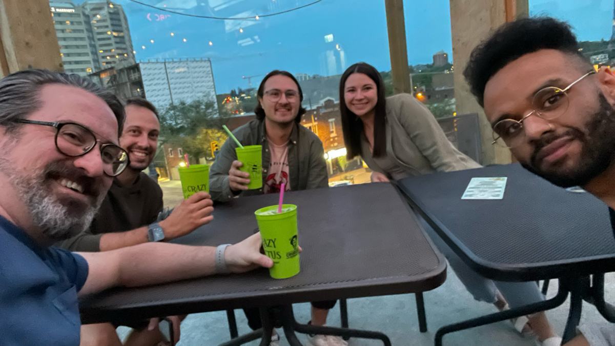 Five people smiling around outdoor patio tables at dusk, with green drinks and a city skyline in the background.