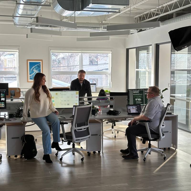 Three colleagues chatting in a bright, open-concept office with multiple computer monitors and large windows.